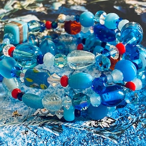 May include: A multi-strand bracelet made of blue, turquoise, and clear glass beads. The beads are of various shapes and sizes, including round, oval, and teardrop.