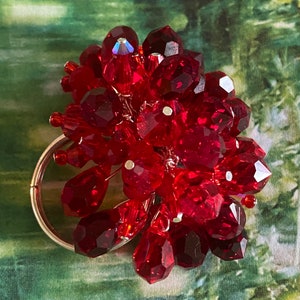 May include: A cluster of faceted red beads, resembling a bunch of grapes, is attached to a silver-colored ring. The beads vary in size and shape, with some teardrop-shaped. The background is a blurred green and white pattern.