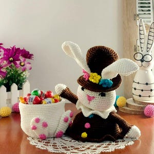 Peut inclure: Un lapin de Pâques au crochet fait à la main, avec un chapeau haut de forme marron et un col blanc. Le lapin tient une tasse blanche remplie de bonbons colorés emballés. La tasse a des pois roses. Une nappe en dentelle blanche est en dessous.
