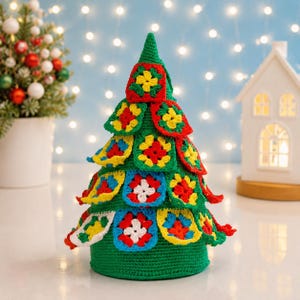 May include: A conical Christmas tree decoration made of green yarn, featuring multiple colorful granny square motifs in red, yellow, blue, and white attached to the exterior. The base is a solid green crocheted cylinder with a flat bottom.