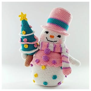 May include: A handmade crochet snowman holding a teal Christmas tree with a yellow star. The snowman wears a pink hat with a teal stripe, a pink scarf, and a pink top with purple and yellow accents. The snowman's body is white with colorful polka dots.