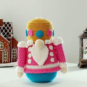 May include: A handmade crochet doll in the shape of a nutcracker. The doll has a yellow head, pink jacket with white accents, and a blue base. The doll is decorated with white buttons and a white beard. The doll is a festive holiday decoration.