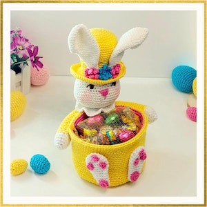 Könnte beinhalten: Ein handgefertigter Häkel-Osterhasenkorb in Gelb, Weiß und Rosa. Der Hase trägt einen gelben Hut mit floralen Akzenten und hält einen Korb voller verpackter Süßigkeiten. Gehäkelte Ostereier in Gelb, Blau und Rosa sind verstreut.