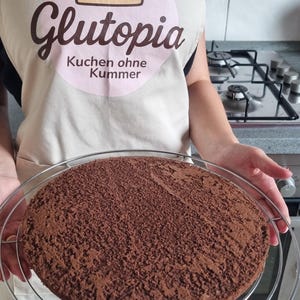Könnte beinhalten: Ein runder Schokoladenkuchen auf einem Metall-Kühlrost. Die Person trägt eine beige Schürze mit dem Text "Glutopia" und "Kuchen ohne Kummer". Die Schürze hat einen rosa Kreis mit einer Kuchenillustration.
