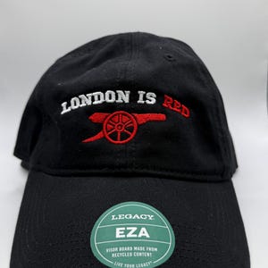 May include: Black baseball cap with the text "LONDON IS RED" embroidered in white and red. The cap features a red cannon logo and a green and white tag that reads "LEGACY EZA".