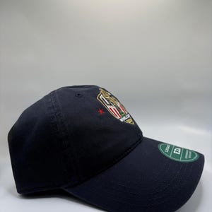 May include: A navy blue baseball cap with a curved brim. The cap features an embroidered emblem with gold, red, and white details. A green tag is attached to the brim.