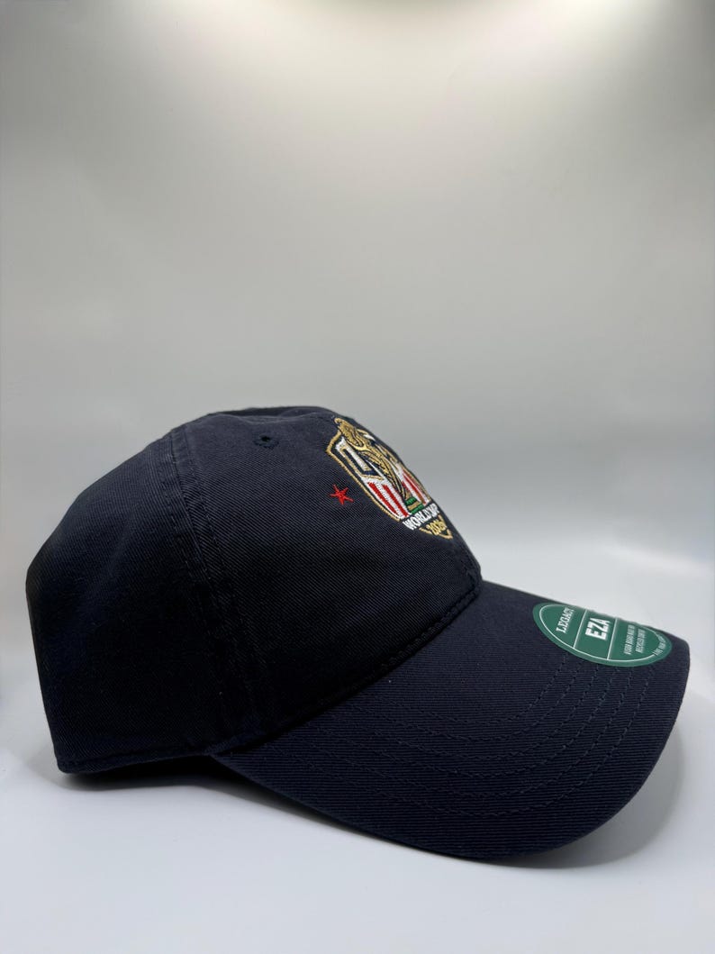 May include: A navy blue baseball cap with a curved brim. The cap features an embroidered emblem with gold, red, and white details. A green tag is attached to the brim.