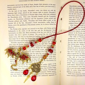 Op de afbeelding: Een ketting met een rode kralenketting en een hanger met een goudkleurige draak en een hartvormige bedel. De hanger heeft rode edelstenen en een goudkleurig zwaard. De ketting wordt getoond op een open boek.
