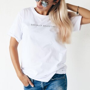 K&ouml;nnte beinhalten: Ein wei&szlig;es kurz&auml;rmeliges T-Shirt mit dem in Schwarz auf die Brust gedruckten Text "SOCIALLY SELECTIVE", kombiniert mit einer blauen Jeans. Das T-Shirt hat einen Rundhalsausschnitt und eine lockere Passform f&uuml;r einen schlichten Look.