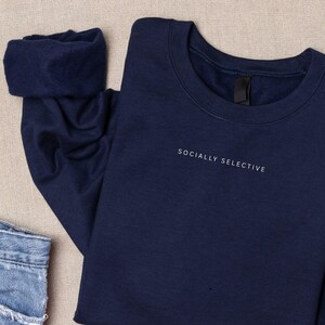 K&ouml;nnte beinhalten: Ein marineblaues Sweatshirt mit Rundhalsausschnitt und dem Text "SOCIALLY SELECTIVE", der in kleinen wei&szlig;en Gro&szlig;buchstaben auf der oberen Brust gestickt ist. Das Kleidungsst&uuml;ck hat lange &Auml;rmel mit gerippten B&uuml;ndchen und einen klassischen Rundhalsausschnitt.