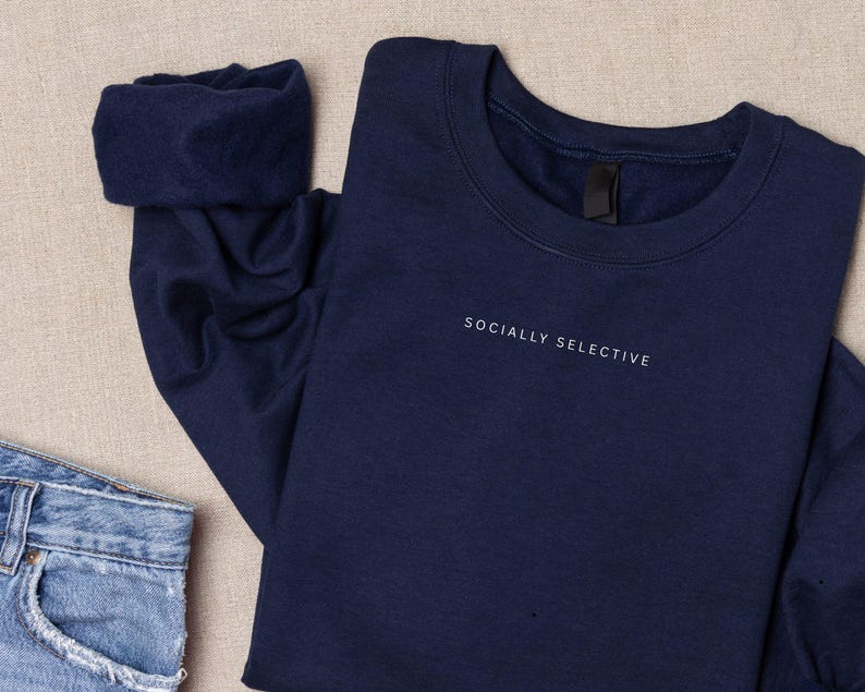 K&ouml;nnte beinhalten: Ein marineblaues Sweatshirt mit Rundhalsausschnitt und dem Text "SOCIALLY SELECTIVE", der in kleinen wei&szlig;en Gro&szlig;buchstaben auf der oberen Brust gestickt ist. Das Kleidungsst&uuml;ck hat lange &Auml;rmel mit gerippten B&uuml;ndchen und einen klassischen Rundhalsausschnitt.