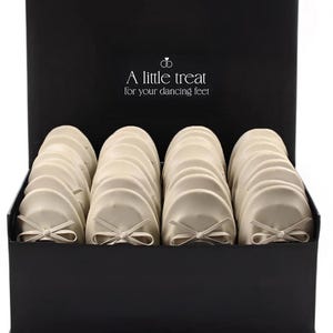 Puede incluir: Una caja rectangular negra que contiene varios pares de bailarinas de color crema, cada una con un pequeño lazo en la parte delantera. El interior de la tapa muestra el texto "A little treat for your dancing feet" con un icono de dos anillos.