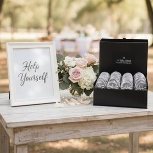 Op de afbeelding: Een witte fotolijst met de tekst Help Yourself, een glazen vaas met roze en witte bloemen, en een zwarte doos met meerdere paren zilveren glitterballerina's met de tekst A little treat for your dancing feet.