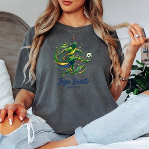 K&ouml;nnte beinhalten: Dunkelgraues T-Shirt mit einer farbenfrohen Grafik eines Fu&szlig;ballspielers, der einen Ball tritt, mit gr&uuml;nen, gelben und blauen Akzenten. Der Text "Joga Bonito Spirit Brazil" ist darunter aufgedruckt. Das Shirt ist aus weichem Material.