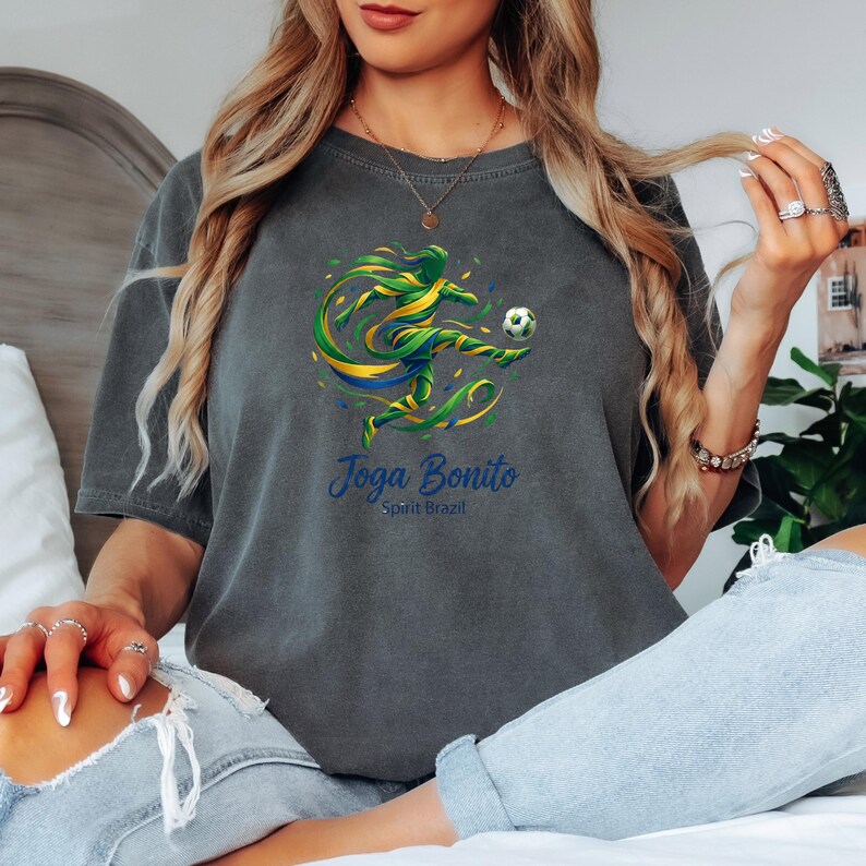 K&ouml;nnte beinhalten: Dunkelgraues T-Shirt mit einer farbenfrohen Grafik eines Fu&szlig;ballspielers, der einen Ball tritt, mit gr&uuml;nen, gelben und blauen Akzenten. Der Text "Joga Bonito Spirit Brazil" ist darunter aufgedruckt. Das Shirt ist aus weichem Material.