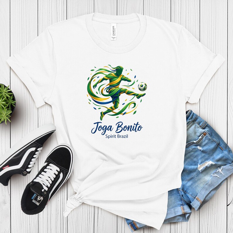 K&ouml;nnte beinhalten: Wei&szlig;es T-Shirt mit einem Fu&szlig;ballspieler-Motiv in Gr&uuml;n, Gelb und Blau, der einen Ball tritt. Der Text "Joga Bonito Spirit Brazil" steht darunter. Schwarz-wei&szlig;e Turnschuhe und Jeansshorts sind ebenfalls zu sehen.