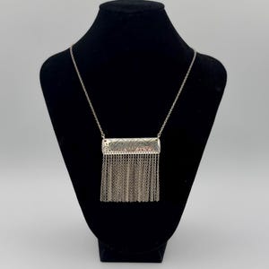 May include: Silver-toned necklace with a rectangular pendant featuring intricate etched designs and a fringe of delicate chains. The necklace is displayed on a black mannequin, showcasing its elegant details and craftsmanship.