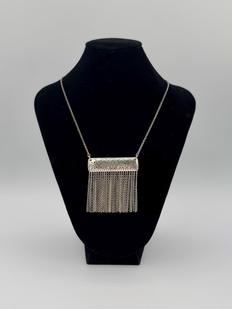 May include: Silver-toned necklace with a rectangular pendant featuring intricate etched designs and a fringe of delicate chains. The necklace is displayed on a black mannequin, showcasing its elegant details and craftsmanship.