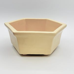Może przedstawiać: Sześciokątna ceramiczna donica w miękkim, kremowo-żółtym kolorze. Wnętrze ma jasny odcień brzoskwini. Donica ma lekko rozszerzający się kształt i stoi na trzech małych nóżkach. Idealna do sukulentów lub małych roślin.