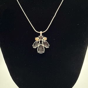 May include: A silver-toned necklace featuring a pendant with a cluster of dark black teardrop and marquise-shaped stones, accented by two smaller light brown round stones at the top, all set in a metallic frame on a thin silver-toned chain.