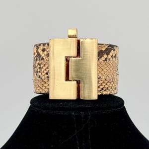 May include: A wide cuff bracelet featuring a tan and brown snakeskin pattern with a prominent, brushed gold-tone metal rectangular clasp centered on the front. The band is constructed with visible zigzag stitching along the edges of the textured material.