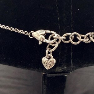 May include: Silver-toned necklace clasp with a heart-shaped charm. The charm has a cursive "B" inside the heart. The necklace chain is visible on either side of the clasp, with a series of small links.