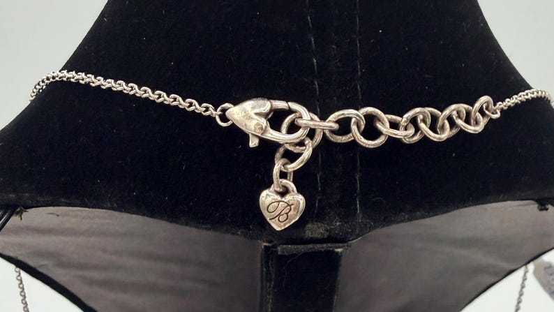 May include: Silver-toned necklace clasp with a heart-shaped charm. The charm has a cursive "B" inside the heart. The necklace chain is visible on either side of the clasp, with a series of small links.