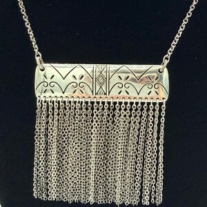 May include: Silver-toned necklace featuring a rectangular pendant with intricate etched patterns and a fringe of delicate chains. The pendant has a geometric design with floral and diamond-shaped elements. The necklace hangs on a fine chain.