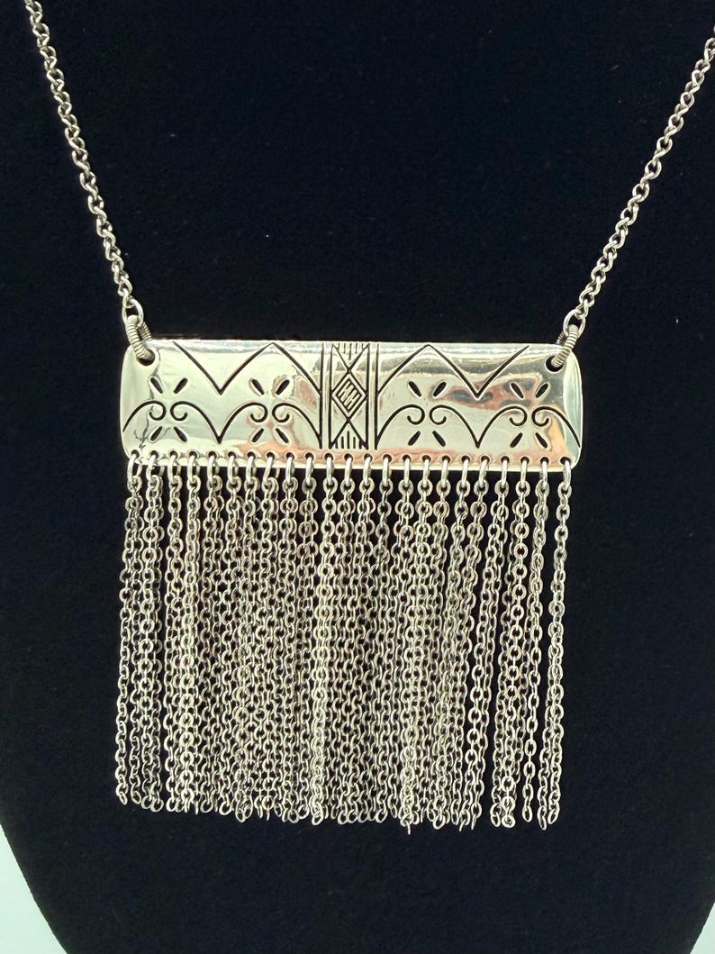 May include: Silver-toned necklace featuring a rectangular pendant with intricate etched patterns and a fringe of delicate chains. The pendant has a geometric design with floral and diamond-shaped elements. The necklace hangs on a fine chain.