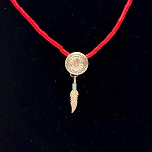 May include: A red beaded necklace with a silver pendant. The pendant has a floral design and a small feather charm. The feather charm is gold-colored and has a small turquoise bead. The necklace is displayed against a black background.