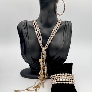 Puede incluir: Un conjunto de joyas con un collar de múltiples hilos con cuentas doradas y una pulsera a juego. El conjunto incluye pendientes de aro grandes. Las joyas se exhiben en una cabeza de maniquí negra y una almohada de terciopelo negro.