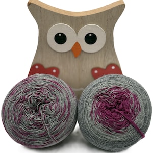 Könnte beinhalten: Zwei Knäuel Garn in Grau- und Magentatönen vor einer Holz-Eulen-Dekoration. Die Eule hat große Augen, einen orangefarbenen Schnabel und rote herzförmige Details. Geeignet zum Stricken oder Häkeln.
