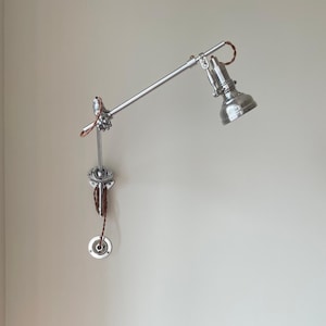 Może przedstawiać: Srebrna, regulowana lampa ścienna z stożkowym kloszem i przegubowym ramieniem. Lampa jest zamontowana na jasnej ścianie nad drewnianym wezgłowiem. Design lampy jest przemysłowy i funkcjonalny.