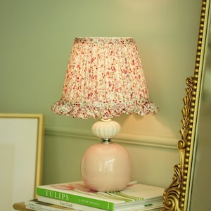 May include: A pink ceramic table lamp with a floral patterned lampshade. The lamp sits on a stack of books, including one titled "TULIPS" by Van Gogh. The lamp has a round base and a ruffled edge on the lampshade.