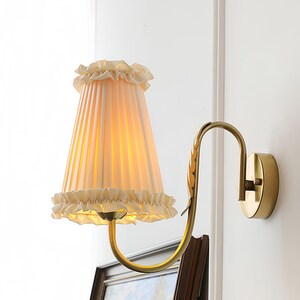 May include: A wall-mounted lamp featuring a brass-toned curved arm with a leaf-shaped detail and a pleated, cream-colored fabric lampshade. The shade is finished with ruffled fabric trim at both the top and bottom edges, emitting a warm glow.