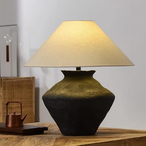 May include: A decorative table lamp with a beige lampshade and a black and gold ceramic base. The lamp is turned on, illuminating the room. The lamp is sitting on a wooden table.