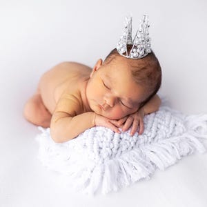 May include: A newborn baby wearing a silver crown with pearl accents, resting on a white, fringed blanket. The baby is sleeping peacefully, with its hands gently supporting its head. The image is set against a bright white background.