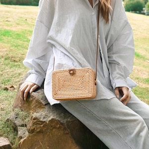 May include: A rectangular, woven straw crossbody bag with a brown leather clasp and strap. The bag is light brown and has a textured surface. The strap is thin and brown. The bag is worn over the shoulder.