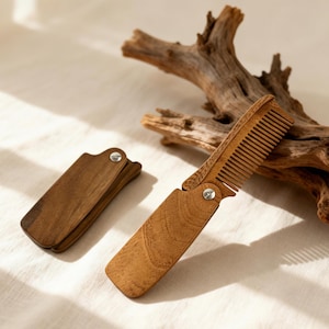 May include: A wooden folding comb, open and closed, with a natural wood grain finish. The comb is attached with a silver rivet. The closed comb is rectangular. The open comb has a handle and teeth. The background includes a piece of driftwood.