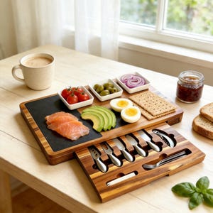 Puede incluir: Una tabla de embutidos rectangular de madera con un cajón extraíble que contiene utensilios de servicio de acero inoxidable. La tabla cuenta con una incrustación de pizarra negra con salmón ahumado, aguacate, huevos duros, galletas y cuencos con aceitunas, tomates y cebolla roja.