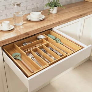 Puede incluir: Un organizador de cajones de bambú con múltiples compartimentos que contiene tenedores, cucharas y cuchillos de acero inoxidable, junto con una espátula de silicona verde claro y una espumadera de silicona verde claro, ordenados en un cajón.