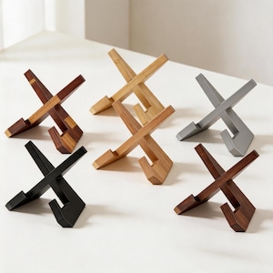 May include: A collection of six wooden and metal phone stands. The stands are in various colors, including dark brown, light brown, black, and silver. Each stand has an X-shaped design with a slot to hold a phone or tablet.