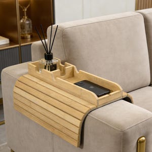 Puede incluir: Una bandeja de brazo de sofá de bambú marrón claro con un soporte para teléfono incorporado y una sección para un difusor de fragancia. La bandeja tiene un diseño curvo para adaptarse al brazo de un sofá. Un teléfono inteligente negro descansa en la bandeja.