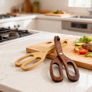 Puede incluir: Dos pinzas de servir de madera, una clara y otra oscura, descansan sobre una encimera cerca de una tabla de cortar de madera con comida. Las pinzas tienen un diseño de tijera con mangos redondeados.