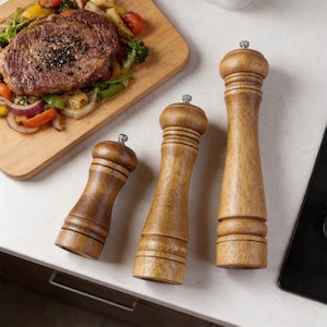 May include: Three wooden pepper grinders of varying sizes. The grinders are made of light brown wood with a classic design. The tops have a small metal cap. A wooden cutting board with a steak and vegetables is in the background.