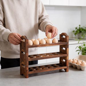 May include: A three-tiered wooden egg holder with a dark brown finish. The holder has a total of 30 circular openings to hold eggs. The image shows a person placing eggs into the top tier. A carton of eggs is visible in the background.