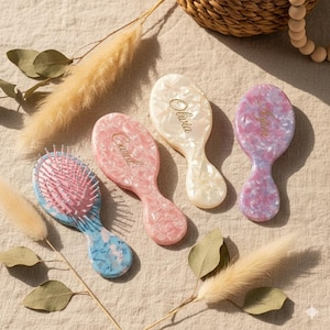 May include: Four personalized hairbrushes in pastel colors. The brushes are oval-shaped with a handle and feature names in gold script: Carol, Olivia, and Aspen. One brush is blue and pink, one is pink, one is white, and one is purple.