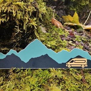 May include: A sticker featuring a mountain range in teal and black, with a yellow camper van silhouette. The sticker is set against a backdrop of moss and foliage, suggesting an outdoor or travel theme.