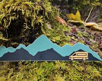 May include: A sticker featuring a mountain range in teal and black, with a yellow camper van silhouette. The sticker is set against a backdrop of moss and foliage, suggesting an outdoor or travel theme.