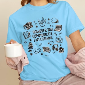 May include: Pale blue t-shirt featuring the phrase "HOWEVER YOU COMMUNICATE, I AM LISTENING." surrounded by black and white illustrations of communication tools. Includes a phone, laptop, and speech bubbles.